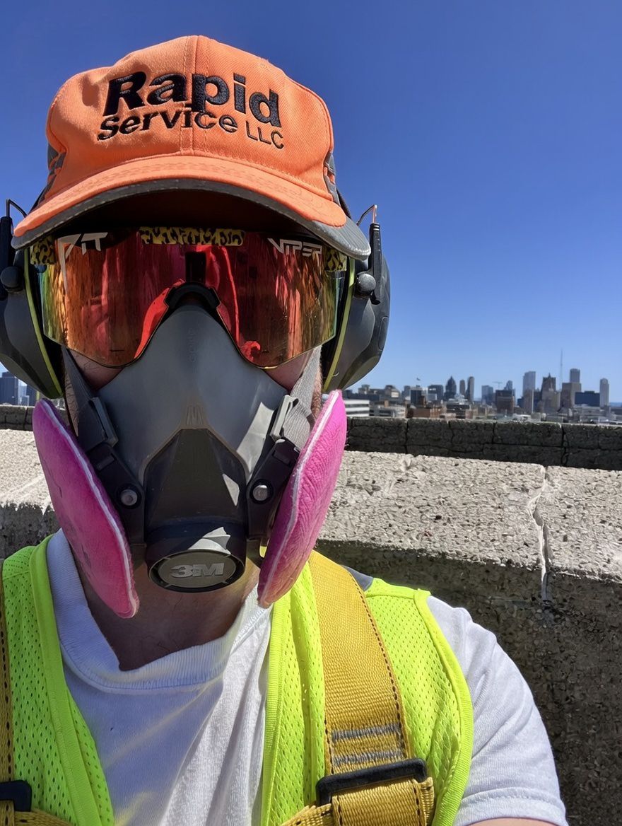 Jacob of Rapid Service L.L.C. on a Milwaukee rooftop in an orange Rapid Service cap, respirator, and hi-vis vest.