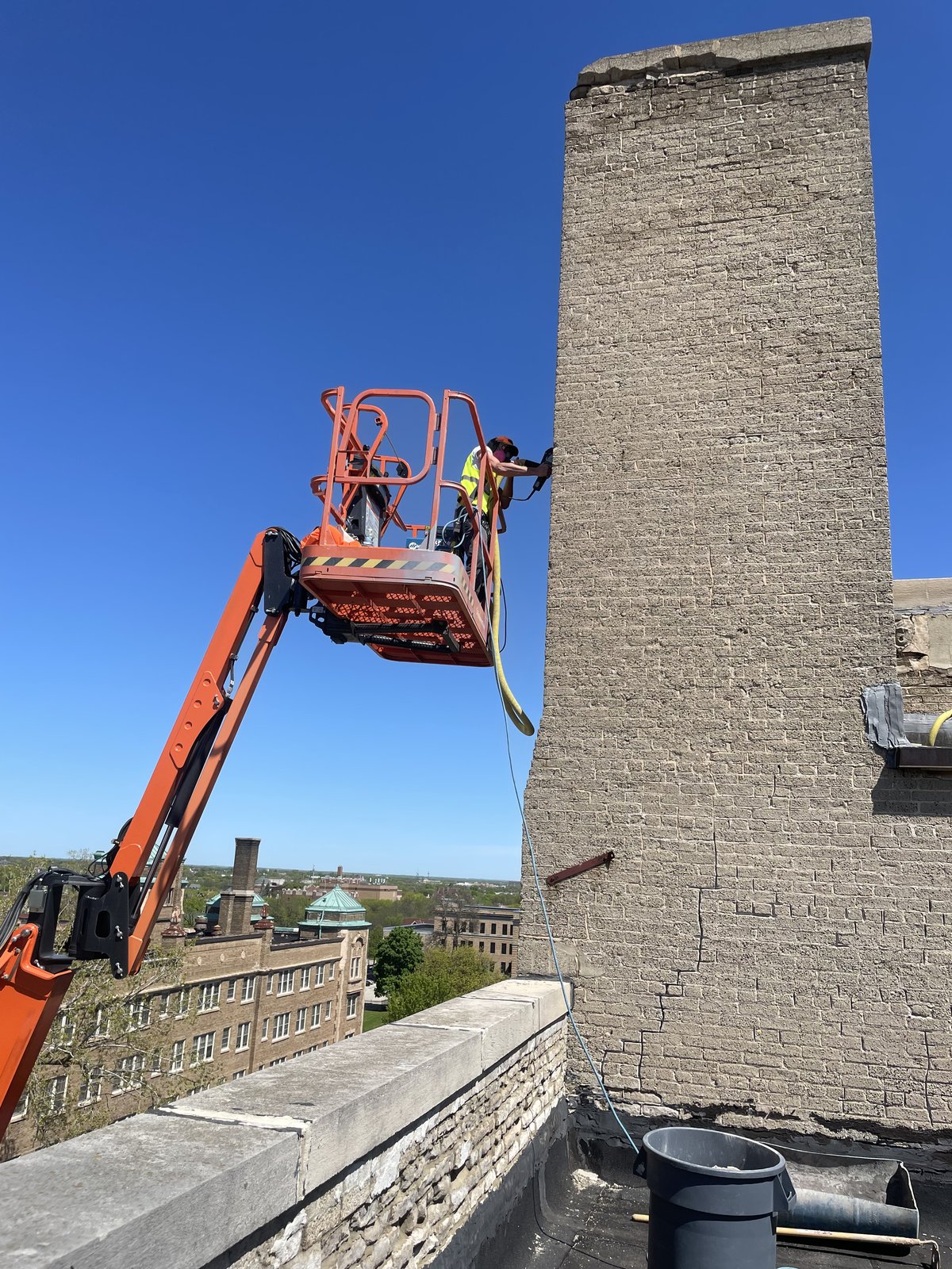 Crew in a boom lift tuckpointing a tall tan brick chimney on a commercial roof.